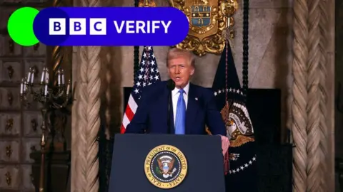 Getty Images President Donald Trump speaks to reporters from a lectern bearing the seal of the US president. An American flag and a flag bearing the president's seal are behind him.  He wears a dark suit and a blue tie.