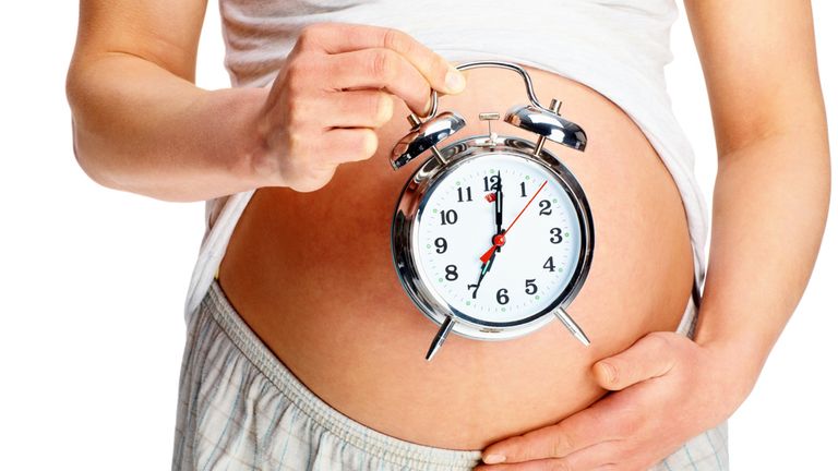 Mid section of a young woman holding an alarm clock against her pregnant belly on white