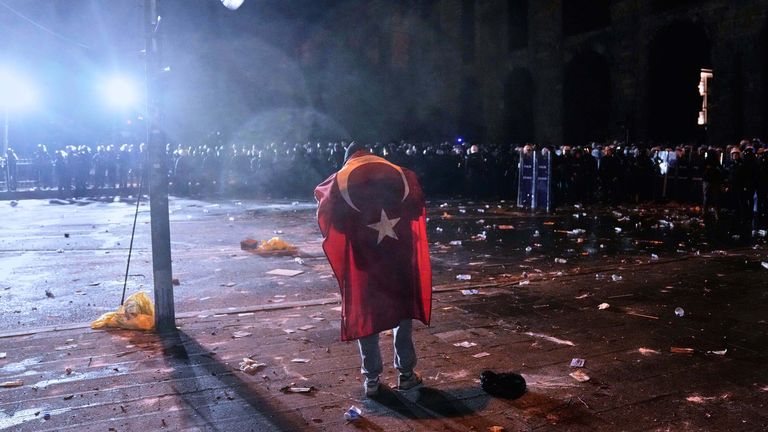 A man, a Turkish flag draped on his back, stands in front of anti riot police officers during clashes in a rally against the arrest of Istanbul's Mayor Ekrem Imamoglu, in Istanbul, Turkey, Friday, March 21, 2025. (AP Photo/Khalil Hamra)